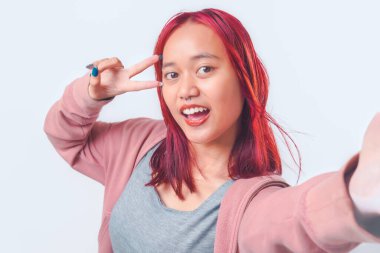 Pembe ceketli güzel genç Asyalı kız beyaz arka planda izole edilmiş iki parmak ya da barış işareti tutarken selfie çekiyor..