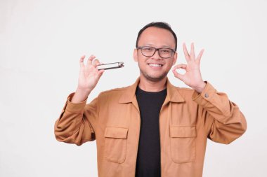 Neşeli genç Asyalı adam, beyaz arka planla izole edilmiş iyi bir jest gösterirken elinde elektrikli sigara tutuyor..