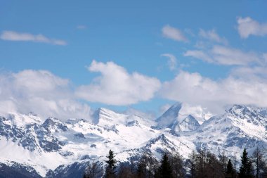 Köknar ağaçlarının tepeleriyle dağlık bir arazi. Kış manzarası, karla kaplı uzak dağlar, bulutlu mavi gökyüzü, güneşli bir gün. İsviçre alplerindeki karlı dağ zirveleri, Wallis kantonu.