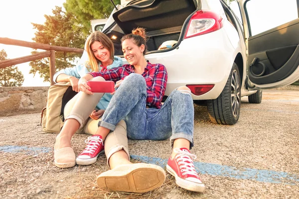 Young female best friends having fun with mobile smart phone at car trip moment - Wanderlust concept during happy travel vacation on the road - Warm late afternoon color tones with main focus on faces — Stock Photo, Image