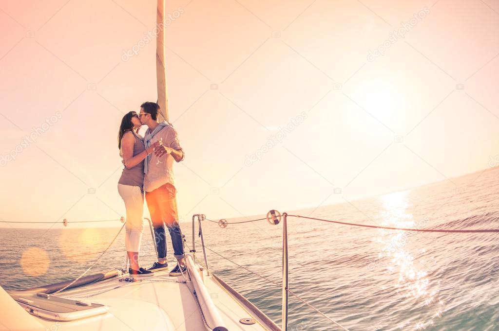 Rich young couple in love on sailboat cheering at sunset - Happy wander lifestyle concept sailing around world - Soft focus on rose quartz filter - Lens flare and tilted horizon as part of composition
