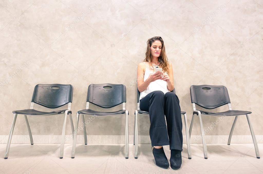 Pensive young woman with mobile smart phone sitting in waiting room - Anxious female person using smartphone in hospital anteroom looking forward for exam test result - Neutral desaturated color tones