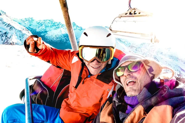 Best friends having fun taking selfie at chairlift with snowboard equipment on mountain ski resort - Winter friendship concept with young people ready to ride down the slope - Bright vivid filter