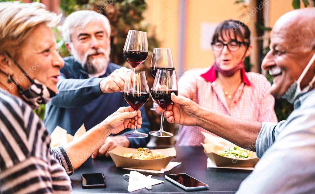 Senior people toasting wine at restaurant bar wearing opened face masks - New normal friendship concept with happy mature friends having fun together at home patio - Warm filter with focus on glasses