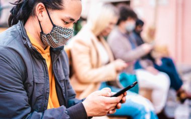 Asyalı adam Covid 'in ikinci dalgasında yüz maskesi takarak akıllı telefon kullanıyor - yeni normal yaşam tarzı kavramı cep telefonundan haber izleyen milenyumlar - göz odaklı sığ alan derinliği