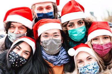 Arkadaşların yüz maskesi ve Noel Baba şapkası takarak selfie çekerken yakın plan fotoğrafı - insanların açık havada eğlendiği yeni normal Noel konsepti - Orta Asyalı adama odaklı parlak filtre