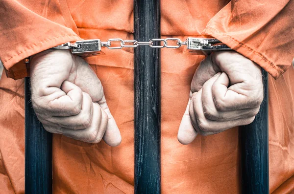 Handcuffed hands of a prisoner behind the bars of a prison with orange clothes - Crispy desaturated dramatic filtered look Royalty Free Stock Photos