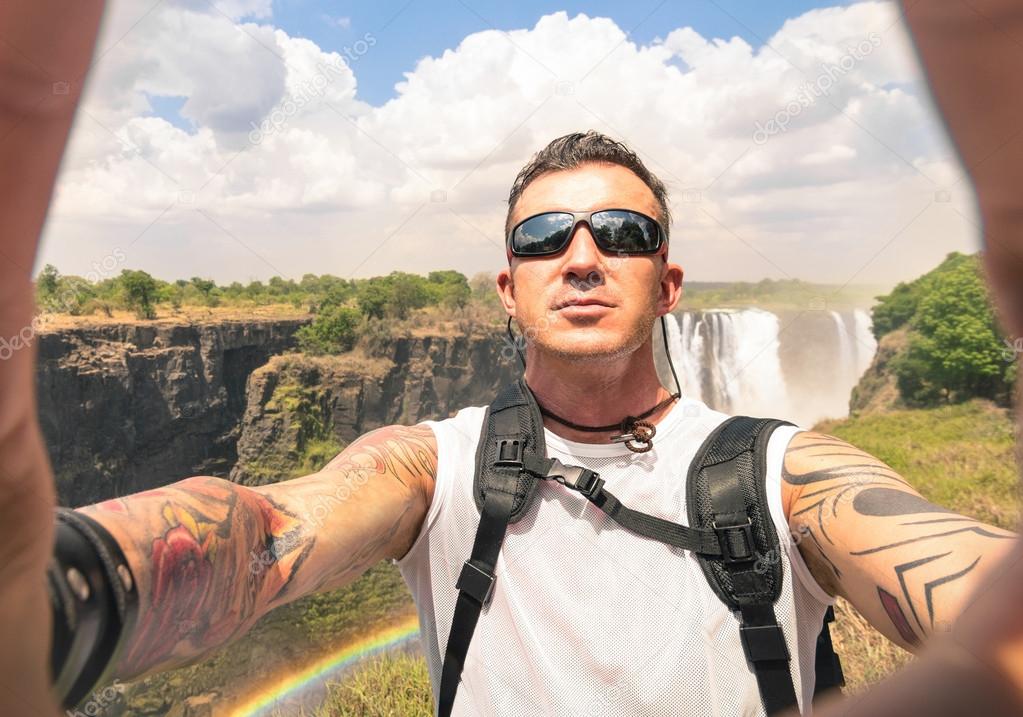 Modern hipster young man taking a selfie at Victoria Waterfalls - Adventure travel lifestyle enjoying moment of connection with nature - Trip excursion in Africa Zimbabwe nature wonder destination