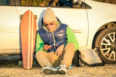 Vintage bir yaşam tarzı - gün batımında sıcak renkler ile genç hipster adam sonraki Kış moda giysileri - modern teknolojileri kavramı arabasıyla oturan smartphone ile karışık