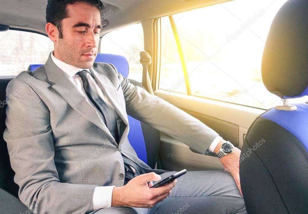 Young handsome businessman sitting in taxi cab while texting sms with smartphone - Business concept with modern man using smart phone - Soft vintage editing with artificial sunlight from the window