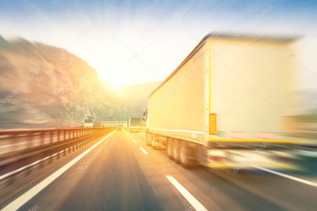 Generic semi trucks speeding on the highway at sunset - Transport industry concept with semitruck containers driving to the mountain pass - Warm editing with pop filtered sunshine and blurred edges