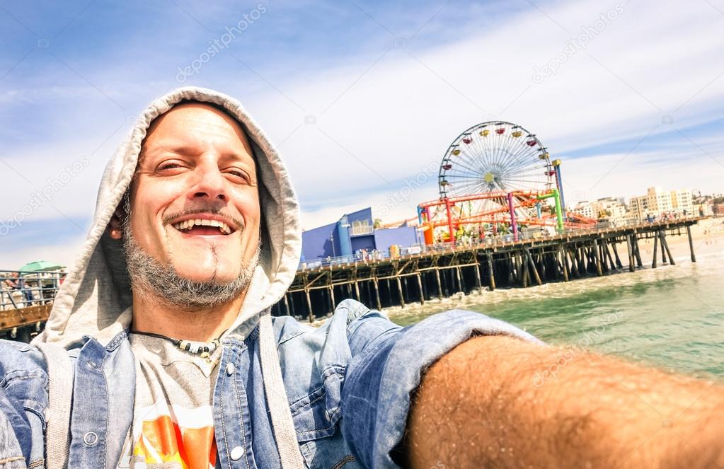 Handsome man taking a selfie at Santa Monica Pier with ferris wheel - Sunny day in California coast - Adventure travel lifestyle around United States of America - Composition with tilted horizon
