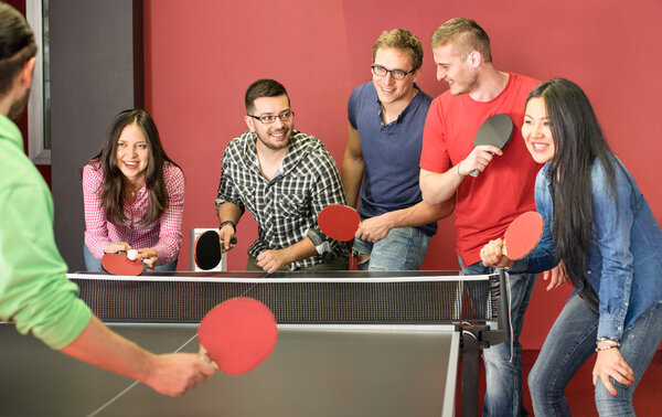 Group of happy young friends playing ping pong table tennis - Fun moment in game room of traveler youth hostel - Concept of vintage sport and genuine emotions - Main focus on two guys with eye glasses