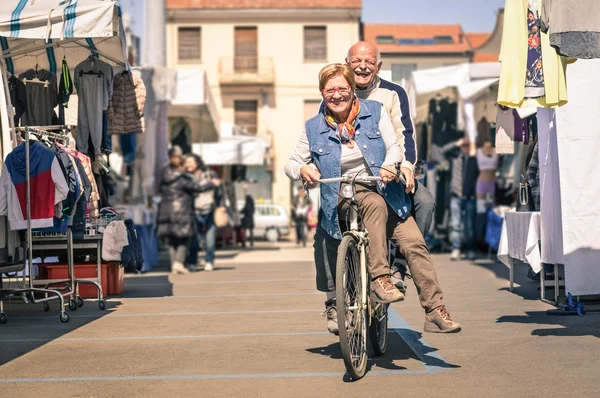 Pareja mayor feliz divirtiéndose con la bicicleta en el mercado de pulgas - Concepto de ancianos activos juguetones con bicicleta durante la jubilación - Estilo de vida de alegría todos los días sin limitación de edad en una tarde soleada de primavera —  Fotos de Stock