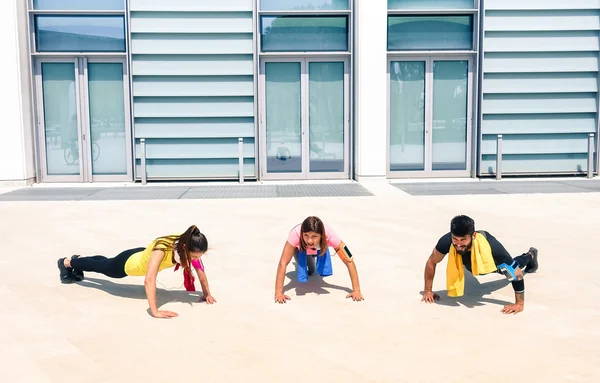 Group of young people performing pushups in modern urban area - Fitness girls exercising with male trainer coach in the city - Sport concept with friends practicing together in a sunny bright day