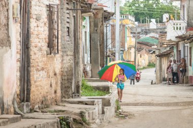 TRINIDAD, CUBA - 22 NOVEMBER, 2015: female kid with multicolored umbrella walking on street in old district side. The town is one of the oldest village founded by Spanish - Unesco World Heritage Site clipart