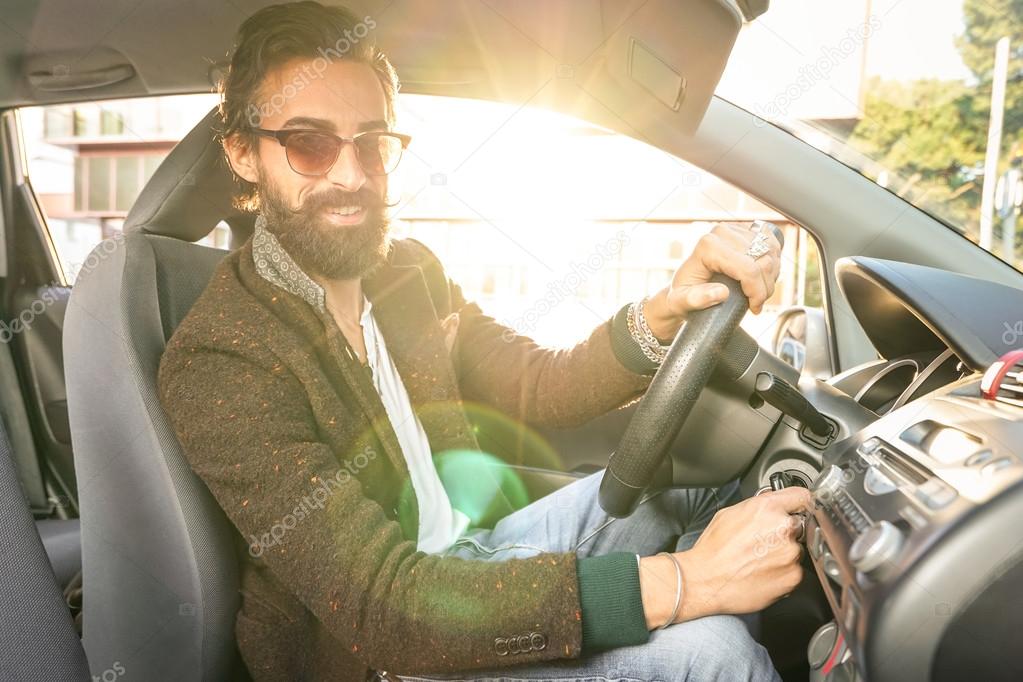 Young hipster fashion model driving car - Young confident man with beard and alternative mustache smiling looking at camera - Warm filter with soft focus on the face due to natural sun flare halo