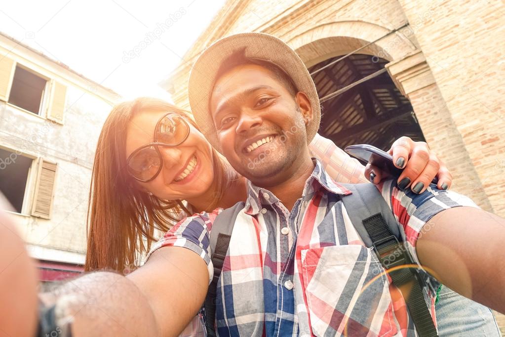 Multiracial couple taking selfie at old town trip - Fun concept with alternative fashion travelers - Indian boyfriend with caucasian girlfriend - Warm filter with powered sunlight and lens flare halo