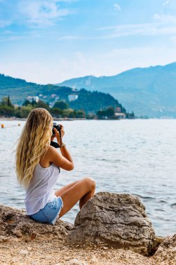Fotoğraf makinesiyle suya yakın oturan kız. Sarışın, dağların fotoğraflarını çekiyor. Arka plan, tatil kavramı