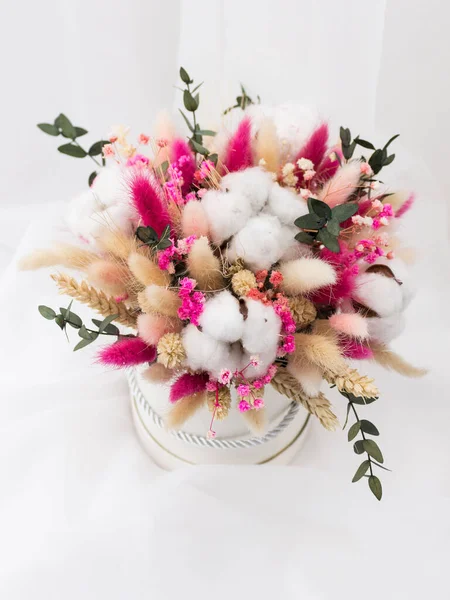 Bouquet of dried flowers in pink, beige, green, white colors in a white cardboard box with a silver rope handle. Top view