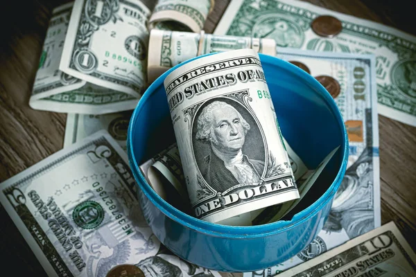 Money, US Dollars ( USD ). An open penny bank shaped like a gas cylinder with one dollar bill on a wooden desk with money scattered on it.