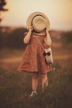 Büyük hasır şapkalı küçük bir kız yüzünü ve kahverengi bir elbiseyle tarlanın arka planında. Romantik kırsal imaj. Yaz batımı. Seçici odak ve tonlama ile resim.