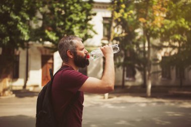 Yakışıklı, sakallı, beyaz saçlı 30 yaşında bir adam sokaktaki plastik şişeden su içiyor. Genç adam, sakallı hippi sıcak yaz gününde suyun tadını çıkar..