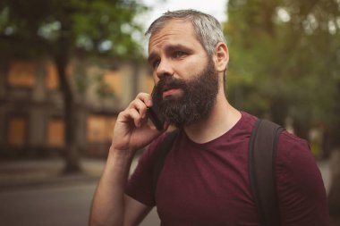 Akıllı telefondan konuşan sakallı genç bir adamın portresi ve şehir caddesinde yürümesi.. 