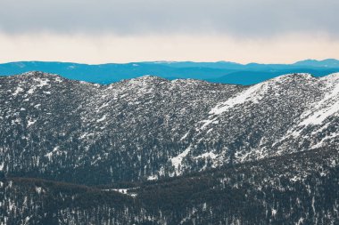 Karla kaplı dağ zirveleri gri bir gökyüzünün altında uzanır, sakin ve geniş bir arazide kışın özünü yakalar.