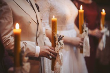 İnsanlar, akşam süslü bir mekanda düzenlenen düğün töreninde kıyafetlerini ve ifadelerini vurgulayan yumuşak ve sıcak bir ışık altında mumlar tutarlar.