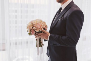 Adam takım elbise giyerken elinde bir buket çiçek tutuyor ve gündüz vakti aydınlık mekanda düğün törenine hazırlanıyor.