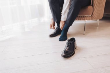 Adam, sabahları aydınlık perdelerle dolu bir odada sandalyede otururken ayakkabı bağcıklarını bağlıyor.