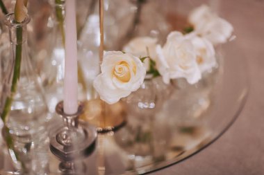 Taze beyaz güller parlak bir masanın üzerinde mumların yanında otururlar. Mumların yumuşak ışığı sıcak bir atmosfer yaratıyor. Bu sahne kapalı bir yerde hazırlandı. Kutlama ya da toplantı için mükemmel.