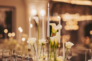 Masada cam vazolara yerleştirilmiş uzun mumlar ve beyaz çiçekler var. Bir ayna yumuşak ışığı yansıtır ve akşam yemeği sırasında sıcak bir atmosfer yaratır.