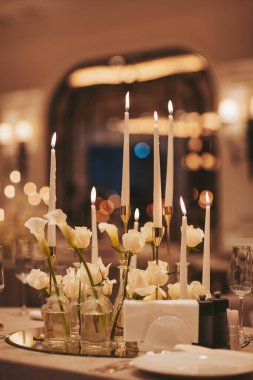 Masada cam vazolara yerleştirilmiş uzun mumlar ve beyaz çiçekler var. Bir ayna yumuşak ışığı yansıtır ve akşam yemeği sırasında sıcak bir atmosfer yaratır.