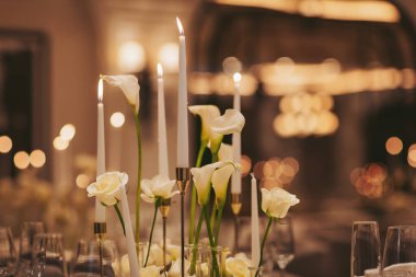 Akşam yemeği için bir yemek masası hazırlanır. Beyaz çiçekler mumların arasında ortada otururlar. Cam eşyalar ve tabaklar düzgün bir şekilde düzenlenir ve olay için resmi bir görünüm oluşturur.