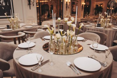 Masalar bir restoranda beyaz çiçekler ve mumlarla kurulur. Yumuşak ışık sıcak bir atmosfer yaratır. Misafirler gece eğlenirken görülebilir.