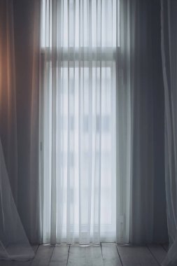 Sessiz bir odadaki perdeler pencereyi kapatır. Işık, yumuşakça hareket eden kumaşın içinden süzülerek ahşap zeminde yumuşak gölgeler oluşturur.