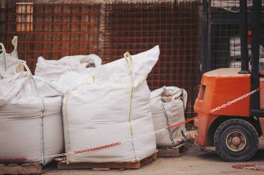 Bir inşaat alanında, paletlerin üzerinde büyük çantaların yanına bir forklift park edilmiştir. İşçiler forklifti malzeme taşımak için kullanabilirler. Ayarlar yoğun ve devam eden inşaat faaliyetlerine odaklanmış durumda.