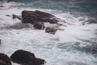 Dalgalar kayalık kıyı şeridine vururken su havaya sıçrıyor. Sahne bulutlu bir gökyüzünün altında engebeli bir kıyı şeridini gösteriyor. Suyun hareketi bir hareket ve enerji hissi yaratır.
