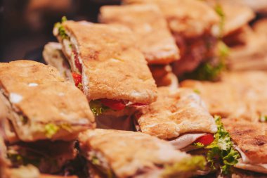 Bir gıda pazarında farklı türde sandviçler bir masaya istiflenir. Sandviçler marul ve domatesli ekmekten yapılır. İnsanlar yoğun bir öğleden sonra yiyecek seçerken görülürler.