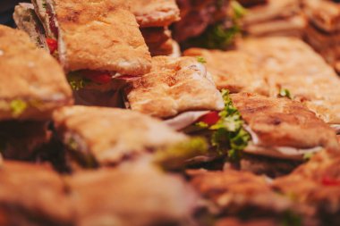 Bir gıda pazarında farklı türde sandviçler bir masaya istiflenir. Sandviçler marul ve domatesli ekmekten yapılır. İnsanlar yoğun bir öğleden sonra yiyecek seçerken görülürler.