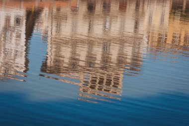 Farklı mimarisi olan bir bina suya yansıyor. Mavi gökyüzü sahnenin üzerinde görünür, suyun yakınındaki yapının genel görüntüsüne eklenir