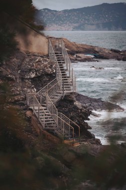 Kırsal metal basamaklar engebeli bir uçurumdan aşağı yuvarlanarak maceraperestleri aşağıdaki çarpışan dalgalara doğru yönlendiriyor. Sahne, doğanın güçlü güzelliğini ve akşam ışığının dingin anlarını yakalar.