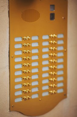 Birden fazla parlak düğmesi olan parlak, altın renkli dahili iletişim paneli sıcak renkli bir duvarda duruyor. Ziyaretçileri davet ederek, modern binanın girişine biraz zarafet katıyor.