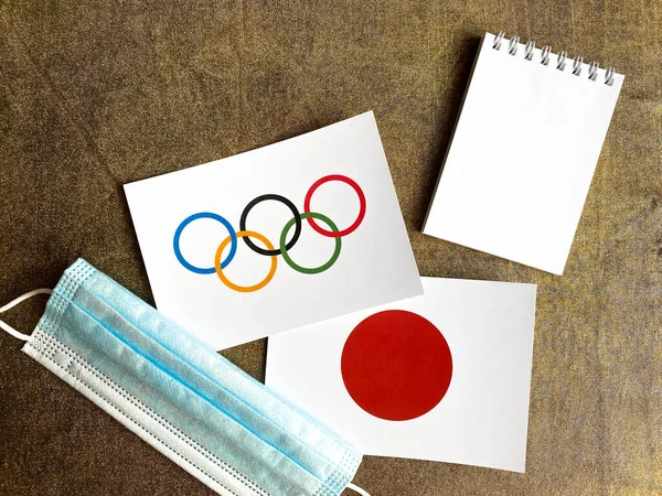 ロシア エル2021年7月22日 オリンピックの旗と日本の国旗が金色に輝く 青い医療マスク 白い空のメモ帳 平置きだ — ストック写真