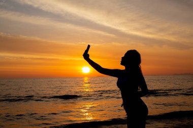 Siluet kadın gün batımında sahilde cep telefonuyla fotoğraf çekiyor..