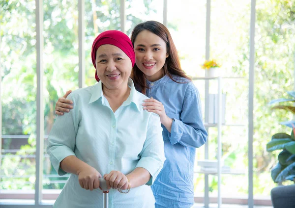 Portrait of Asian elderly mother wearing red headscarf recover from cancer standing, holding walking stick and having her daughter by her side embrace, smile very happily. Cancer or leukemia survivor concept.
