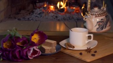 - Çay. Şeffaf koruyucu eldivendeki bir el çaydanlıktan bir fincana çay dökerek şöminenin arka planında çiçekler ve şekerlemeler olan bir masaya koyar..