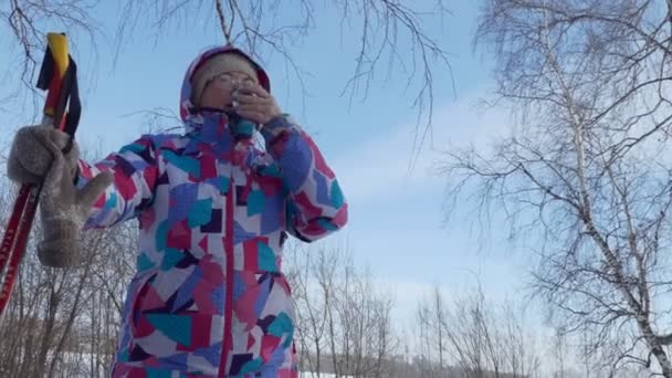 滑雪旅行 一位手里拿着滑雪杆子的老妇人 在森林的树枝和天空的衬托下 用热水瓶盖着咖啡 — 图库视频影像